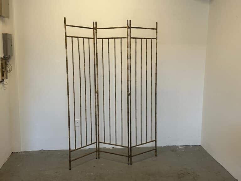 The Maison Bagues Gilt Bamboo Metal Folding Screen, featuring three panels, stands in an empty room with plain white walls and a gray floor.