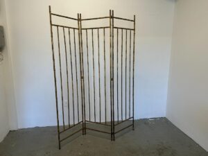A Maison Bagues Gilt Bamboo Metal Folding Screen stands in an empty room with white walls and a gray floor.