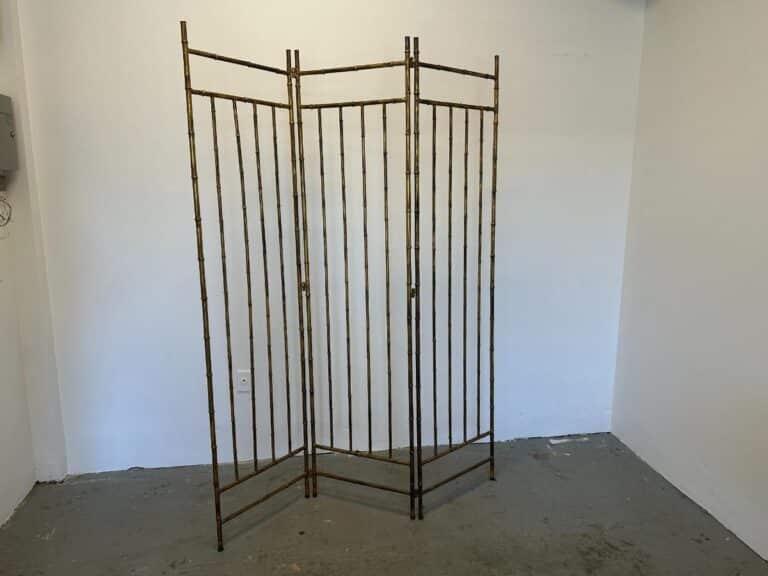 A Maison Bagues Gilt Bamboo Metal Folding Screen stands in an empty room with white walls and a gray floor.