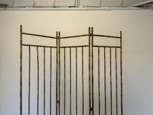A Maison Bagues Gilt Bamboo Metal Folding Screen with three panels stands against a plain white wall.