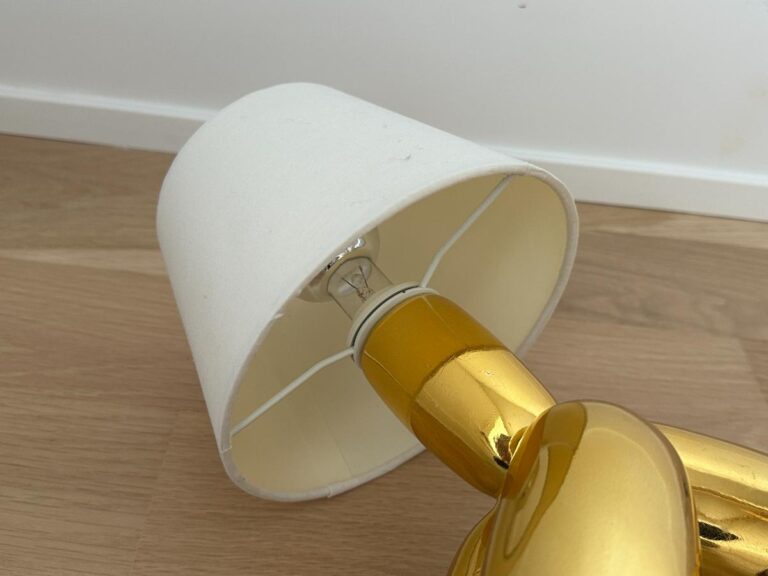 A gold lamp with a white shade lying on its side on a wooden floor near a white wall.