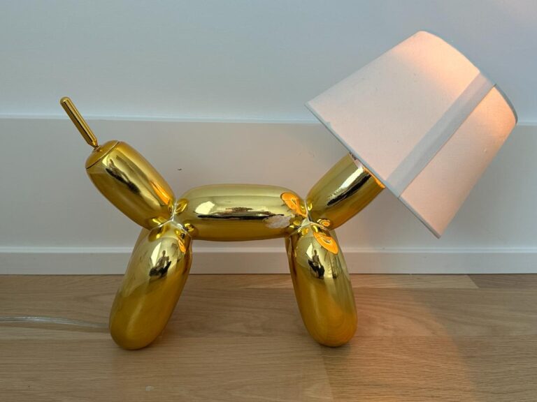 A gold balloon dog sculpture lamp with a white lampshade, placed on a wooden floor.