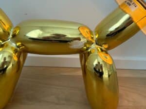 Close-up of a gold balloon dog sculpture with a patch of chipped paint on its side, on a wooden floor.