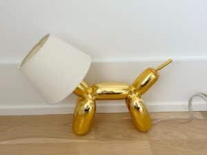A gold balloon dog-shaped lamp with a white lampshade, sitting on a wooden floor against a white wall.