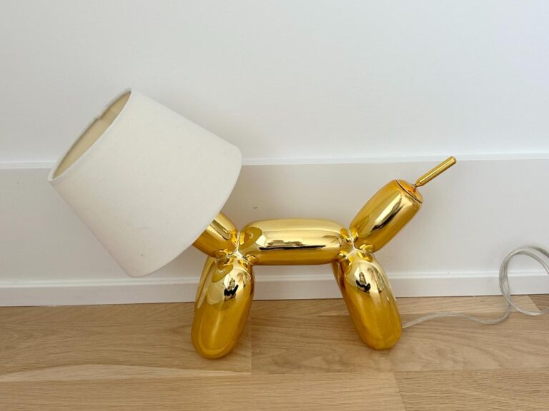 A gold balloon dog-shaped lamp with a white lampshade, sitting on a wooden floor against a white wall.