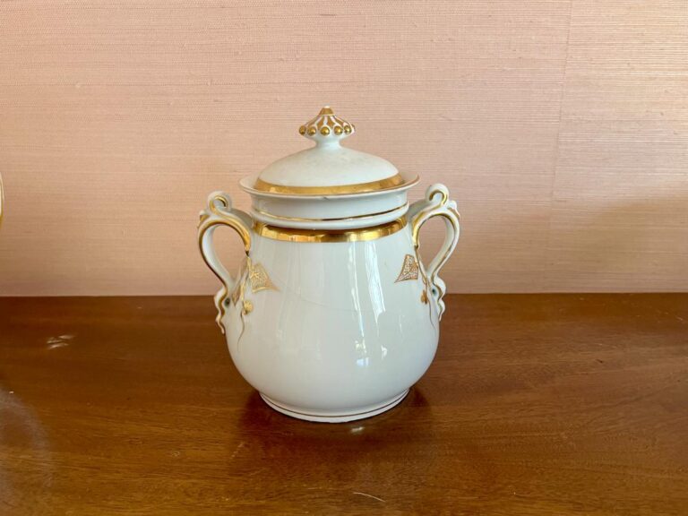 Ornate white ceramic sugar bowl with gold accents and two handles on a wooden surface.