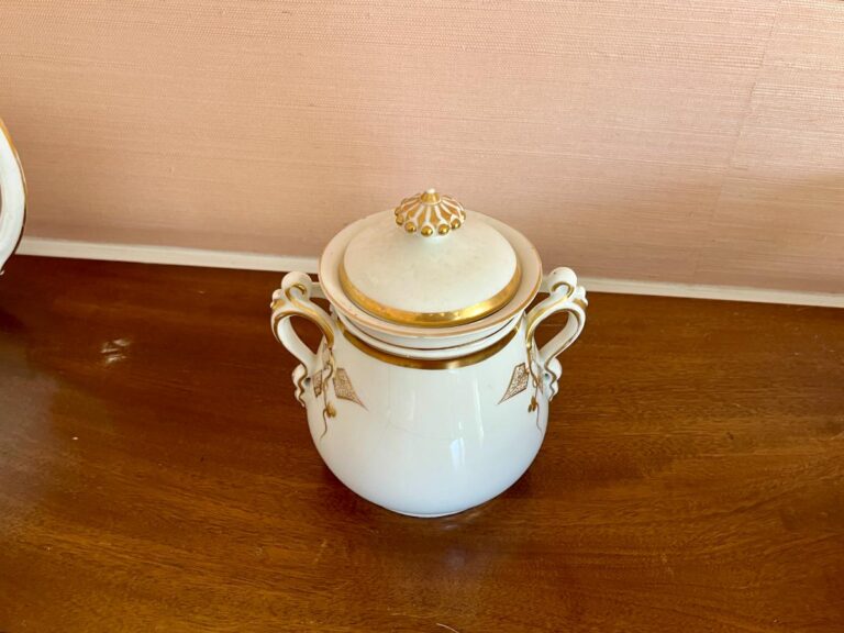 Ornate white and gold ceramic jar with a lid, featuring two handles, on a wooden surface.