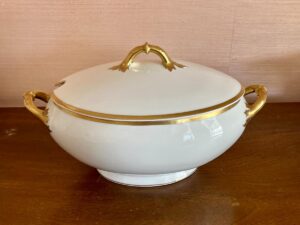 White porcelain tureen with gold trim and handles on a wooden table.