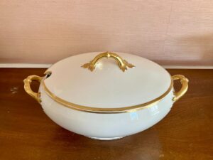 White ceramic serving dish with lid and gold details on handles, displayed on a wooden surface.