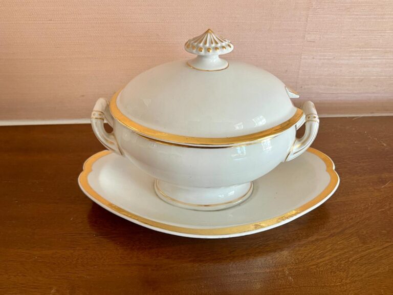 A white porcelain soup tureen with gold trim on a matching plate, placed on a wooden surface.