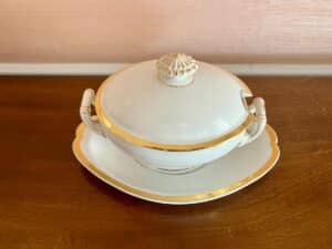 A white porcelain soup tureen with gold trim, lid, and platter on a wooden surface.