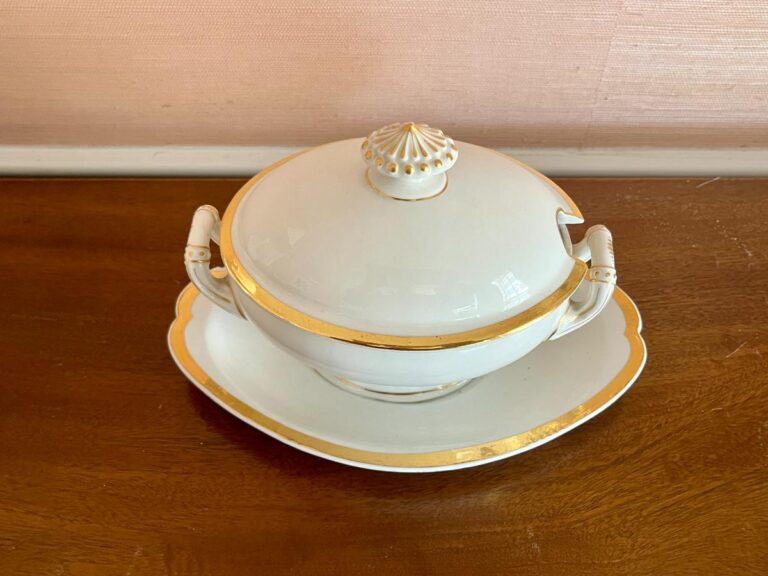 A white porcelain soup tureen with gold trim, lid, and platter on a wooden surface.
