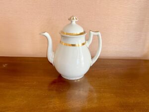 White porcelain teapot with gold accents on a wooden surface, against a light brown textured wall.