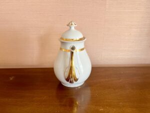 White ceramic teapot with gold accents and a lid on a wooden surface against a beige wall.