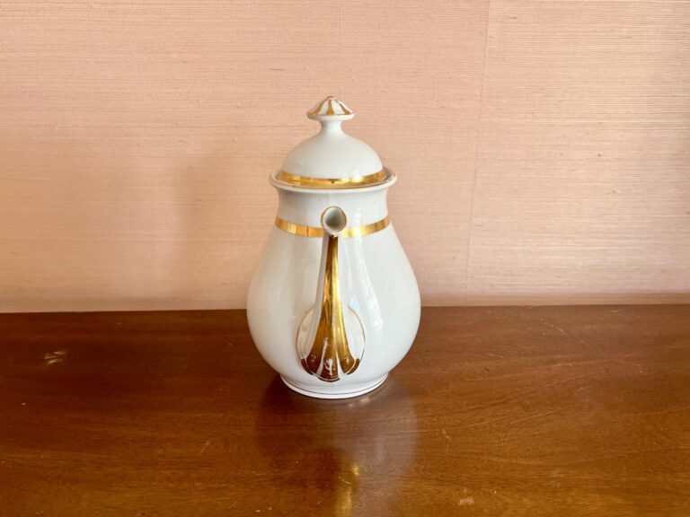 White ceramic teapot with gold accents and a lid on a wooden surface against a beige wall.