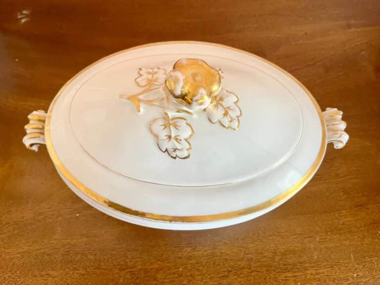 Ornate white ceramic dish with gold accents, floral design on lid, and decorative handles, placed on a wooden surface.