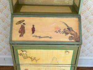 Antique cabinet with Asian art featuring two figures, one with an umbrella, and floral motifs on a painted drawer.