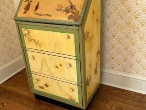 A vintage painted cabinet with mountain and bird designs, set on a wooden floor with patterned wallpaper background.