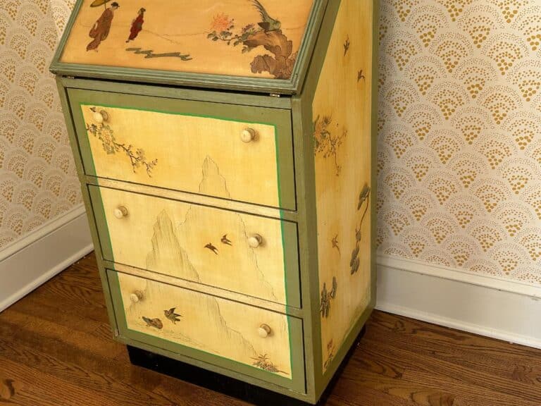 A vintage painted cabinet with mountain and bird designs, set on a wooden floor with patterned wallpaper background.