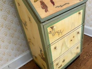 Vintage chest with painted nature scenes and figures on top, in a room with patterned wallpaper and wood floor.