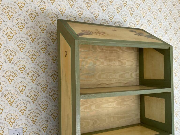 Wooden bookshelf with green trim and nature motifs, set against a patterned wallpaper background.