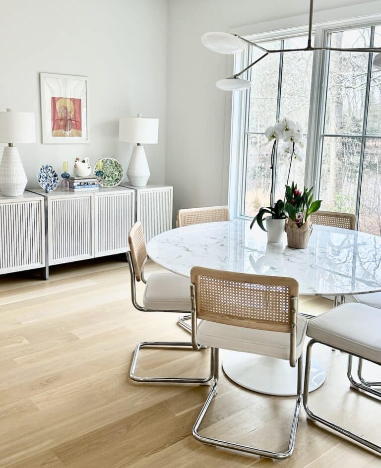 Bright dining room with a marble table, cane chairs, sideboard, lamps, and large windows letting in natural light.