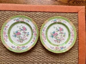 Two decorative plates with floral and bird designs on a woven mat with an orange border.