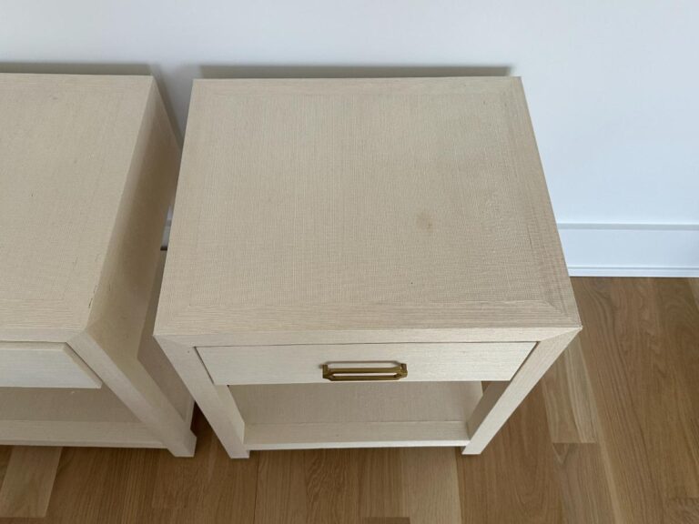 A light-colored wooden nightstand with a drawer and brass handle, on a wood floor next to a white wall.