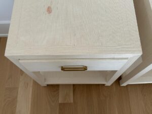 A beige nightstand with a brass handle, showing a reddish stain on the top, on a wooden floor.