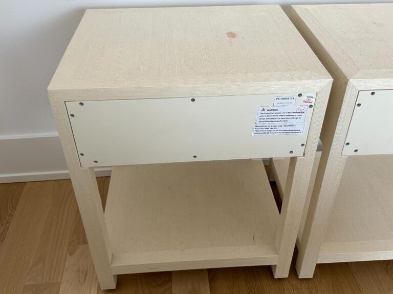 A light wood nightstand with a white panel and warning label on the back, standing on a wooden floor.