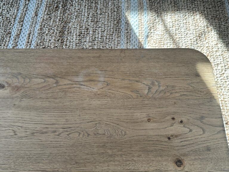 A wooden table with a circular water stain, placed on a textured woven rug with sunlight casting shadows.