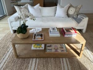 A wooden coffee table with books and a plant sits on a woven rug in front of a white sofa with pillows.