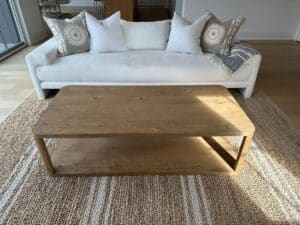 A wooden coffee table on a woven rug in front of a white sofa with patterned and plain pillows.