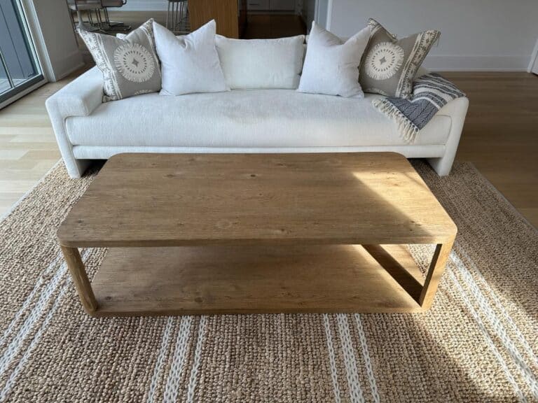 A wooden coffee table on a woven rug in front of a white sofa with patterned and plain pillows.