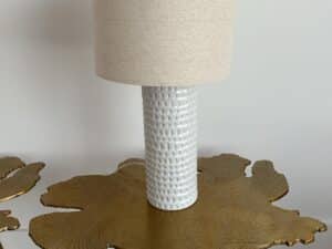 White textured ceramic lamp with a beige shade on a gold table with a leaf-like pattern.