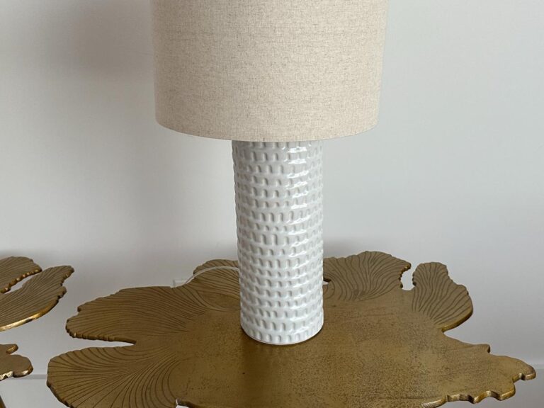 White textured ceramic lamp with a beige shade on a gold table with a leaf-like pattern.