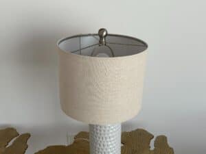 A white textured lamp with a beige lampshade on a table against a plain wall.