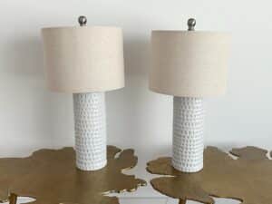 Two white textured table lamps with beige shades on gold leaf-shaped tables against a plain wall.