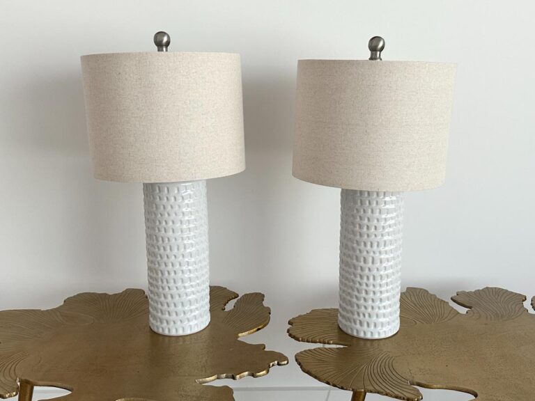 Two white textured table lamps with beige shades on gold leaf-shaped tables against a plain wall.