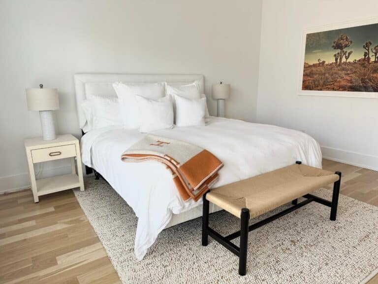 A modern bedroom with a white bed, orange blanket, nightstands, lamps, and a bench at the foot of the bed.