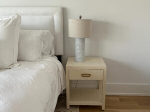 Beige nightstand with a lamp beside a bed with white bedding against a white wall.