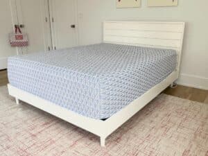 A neatly made bed with a light blue patterned sheet in a bright, minimal bedroom with white walls.