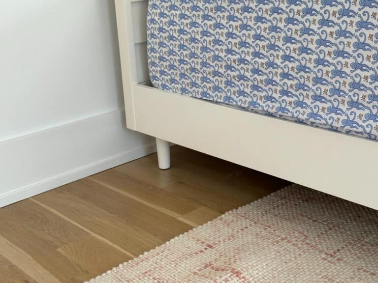 A corner of a white bed with a patterned sheet on a wooden floor and a light-colored rug.
