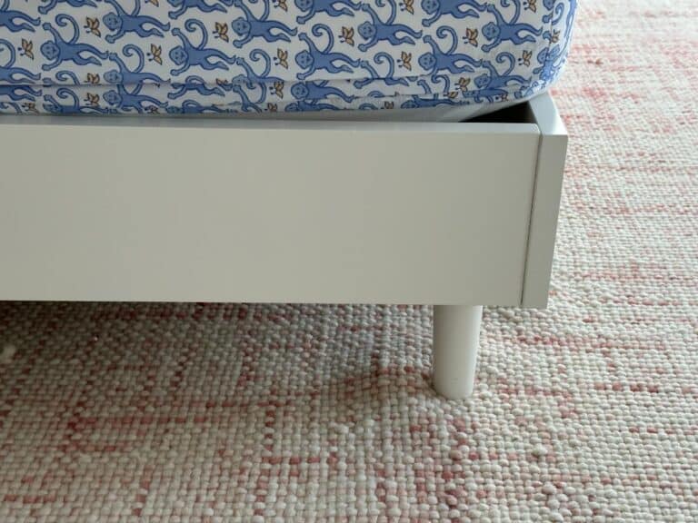 Closeup of a white bed frame with a blue-patterned mattress on a pink and white textured rug.