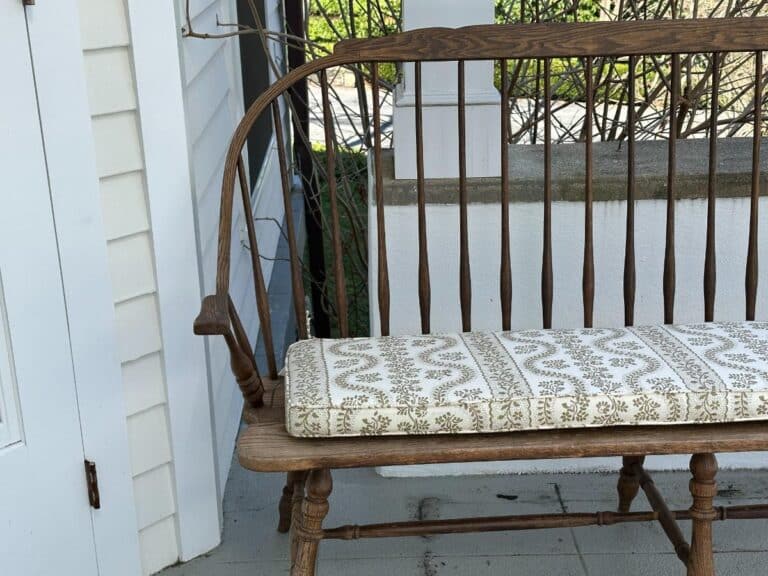 Wooden bench with a patterned cushion on a porch, next to a white wall and greenery.