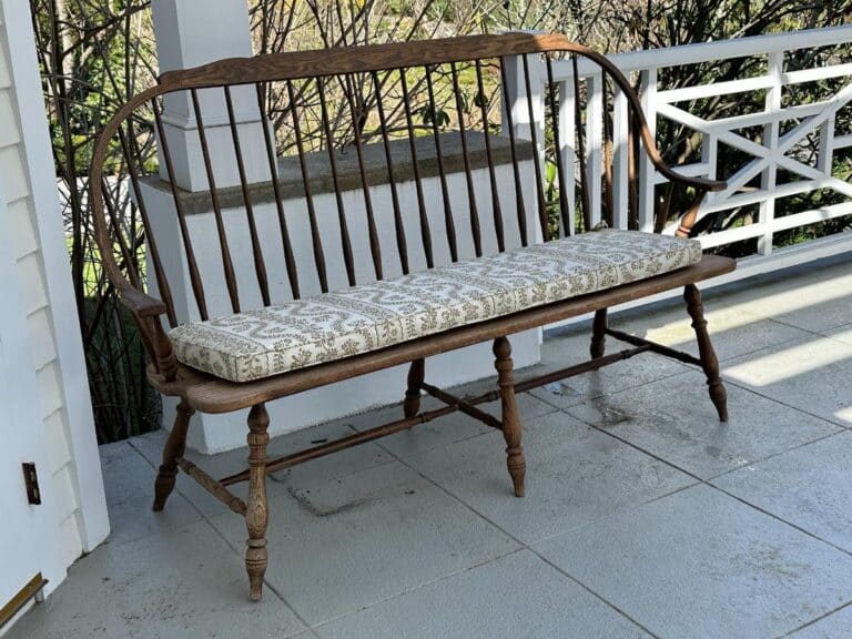Wooden bench with patterned cushion on a porch, surrounded by white railings and trees in the background.
