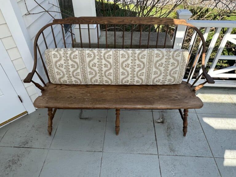 Wooden bench with a patterned cushion on a porch, next to a white door and railing.