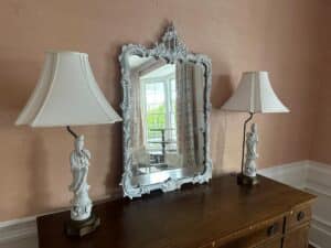 A wooden dresser with a pair of Blanc De Chine Figural Lamps and an elegant mirror reflecting a bright room with large windows.