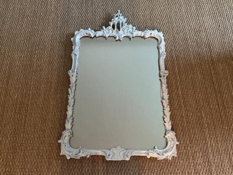 The Vintage Chinoiserie Mirror features an ornate design with a decorative white frame, set against a textured brown surface.
