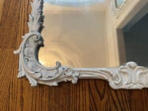 The corner of a room with a Vintage Chinoiserie Mirror, featuring an ornate white frame, reflects the wood floor and arched doorway.